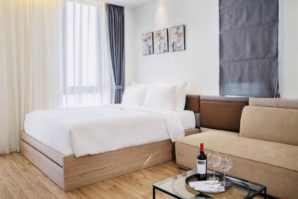 Une chambre à coucher avec un lit, un canapé, une table en verre avec une bouteille et des verres, et une fenêtre avec des rideaux.