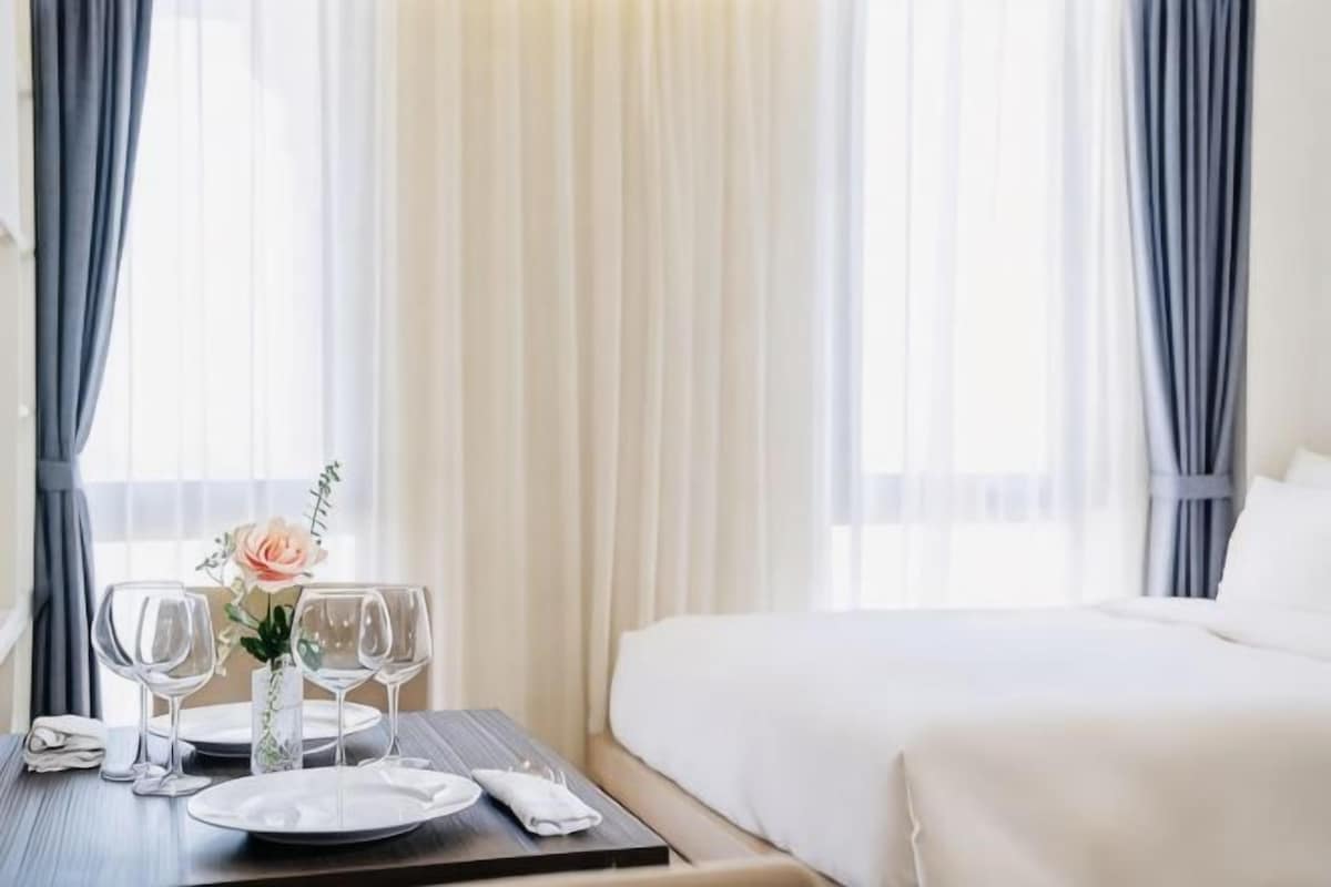 Habitación de hotel con una cama bien hecha, una mesa de comedor con platos y vasos, y un florero con flores.