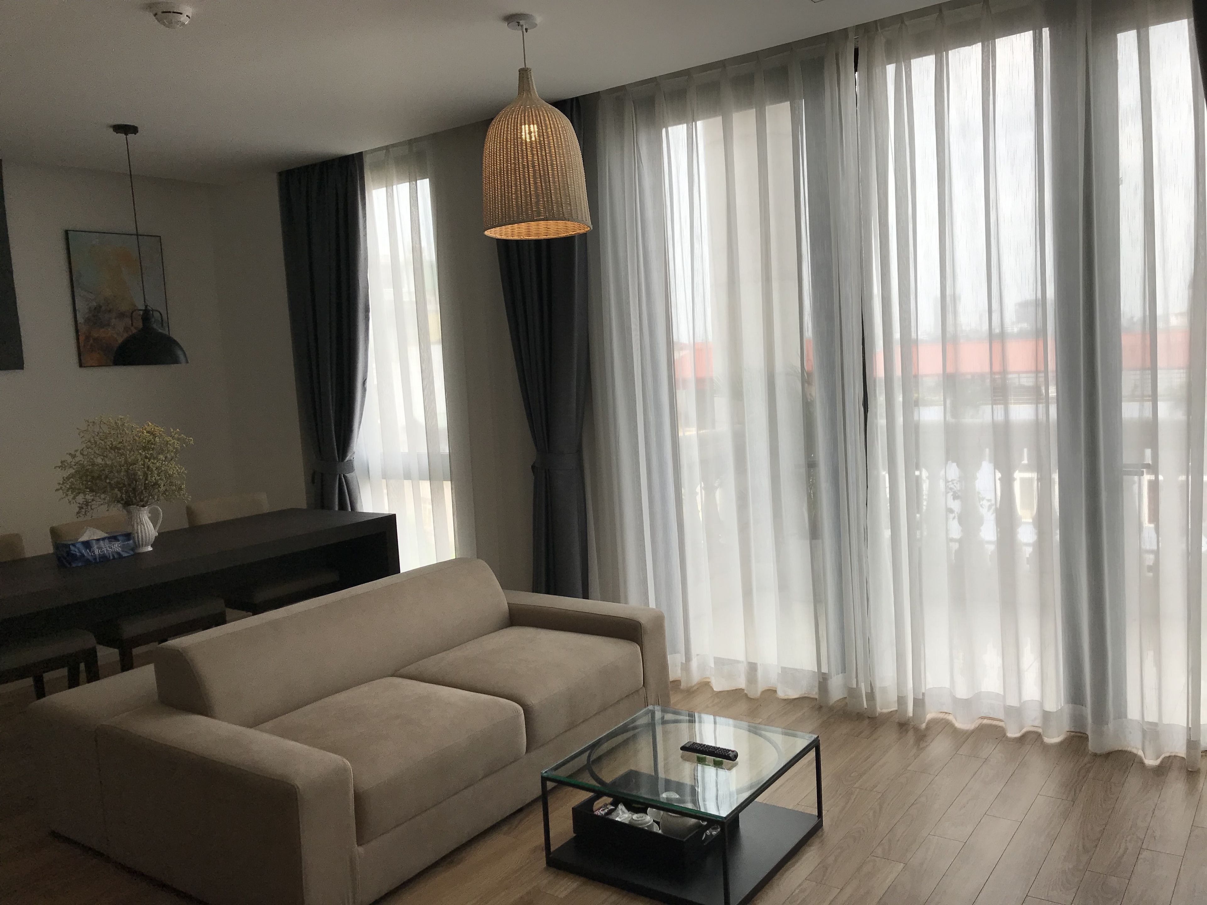 A living room with a sofa, a glass coffee table, a hanging light fixture, and sheer curtains.