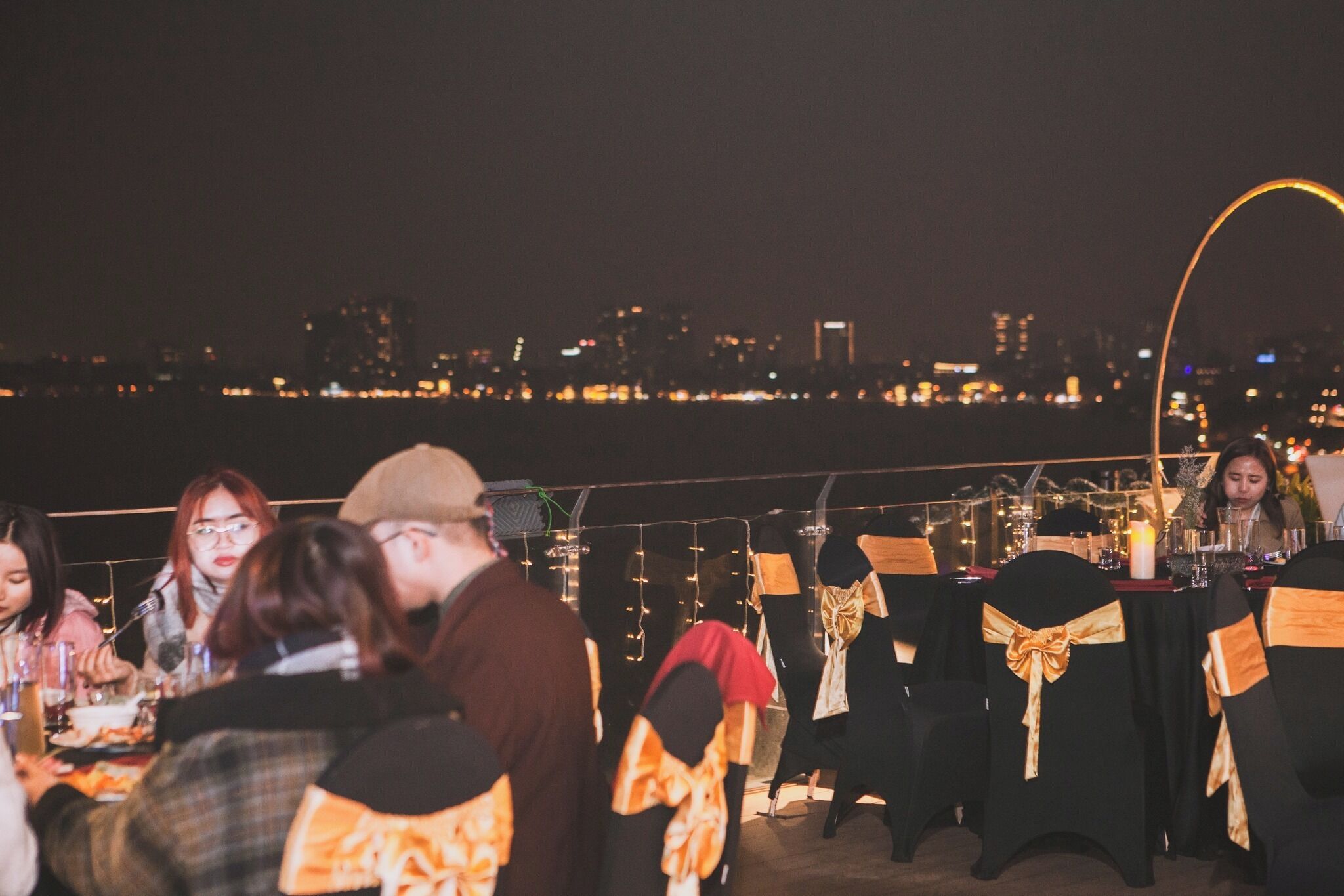 A rooftop dining area with tables set for guests, overlooking a city skyline at night.