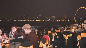 A rooftop dining area with tables set for guests, overlooking a city skyline at night.