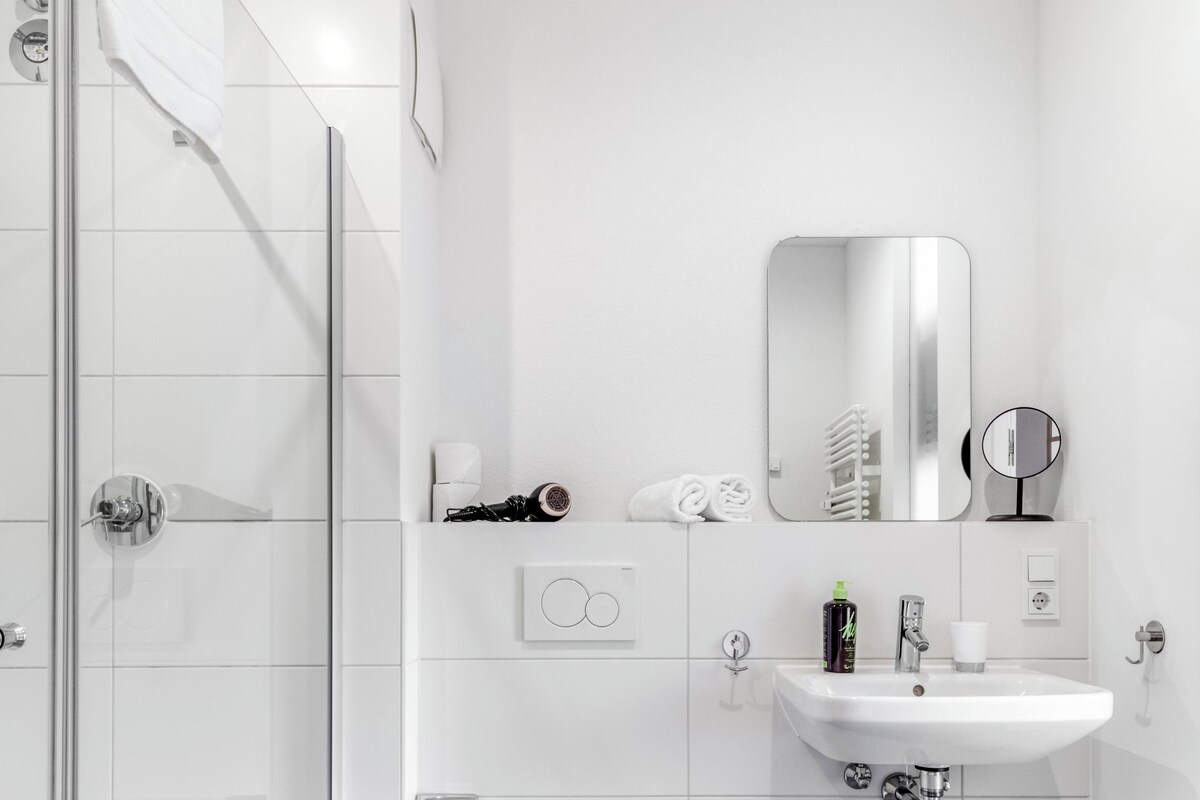 Une salle de bain moderne avec une douche à parois de verre, des toilettes blanches, un lavabo avec un miroir et une poubelle.