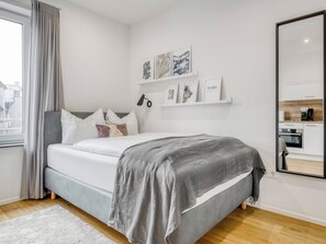 Une chambre à coucher moderne avec un lit, une table de chevet, un miroir et une étagère où sont exposées des photos encadrées.