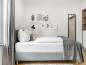 A modern bedroom with a bed, pillows, a bedside table, a mirror, and wall-mounted shelves with framed pictures. - Limehome Augsburg (Augsburg)