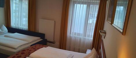 Une chambre d’hôtel avec deux lits, un radiateur, une fenêtre avec des rideaux légers et une lampe fixée au mur.