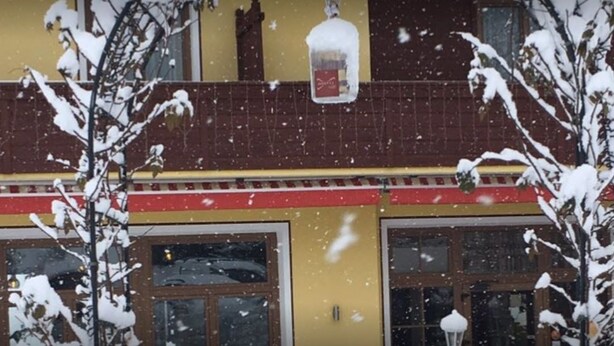 Um edifício com fachada amarela, detalhes em vermelho e grandes janelas. Neve cai lá fora.