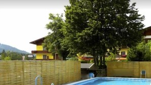 Una piscina rectangular con una escalera metálica, rodeada por una valla de madera, con casas y árboles al fondo.