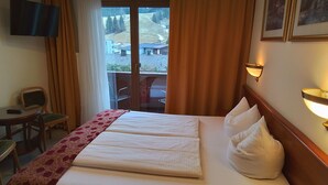Habitación de hotel con cama, escritorio, silla, televisión y vista a un balcón con mobiliario exterior.
