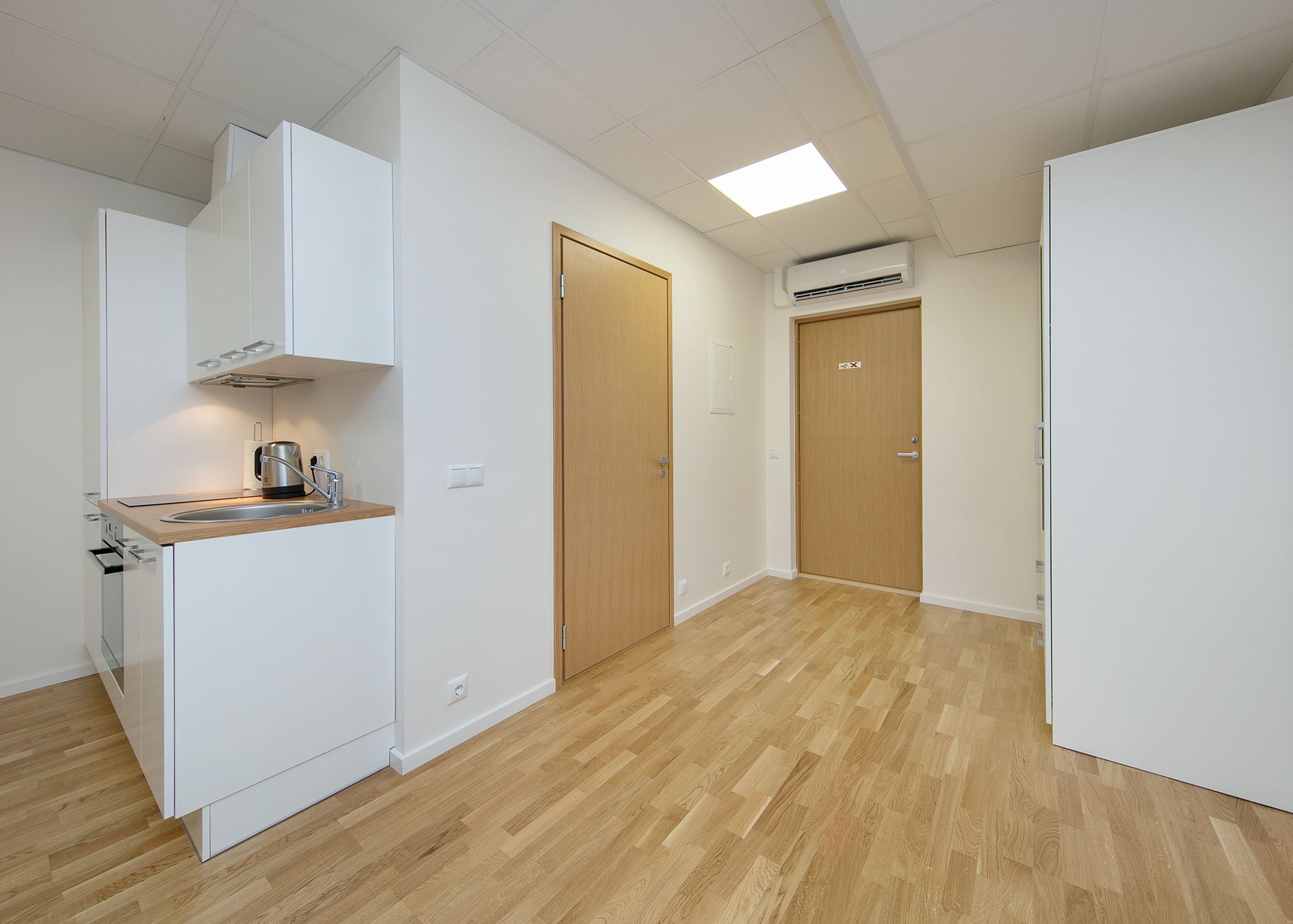 A modern kitchen with white cabinetry, a wooden countertop, and a built-in stove. There is a wooden door and an air conditioning unit on the wall.