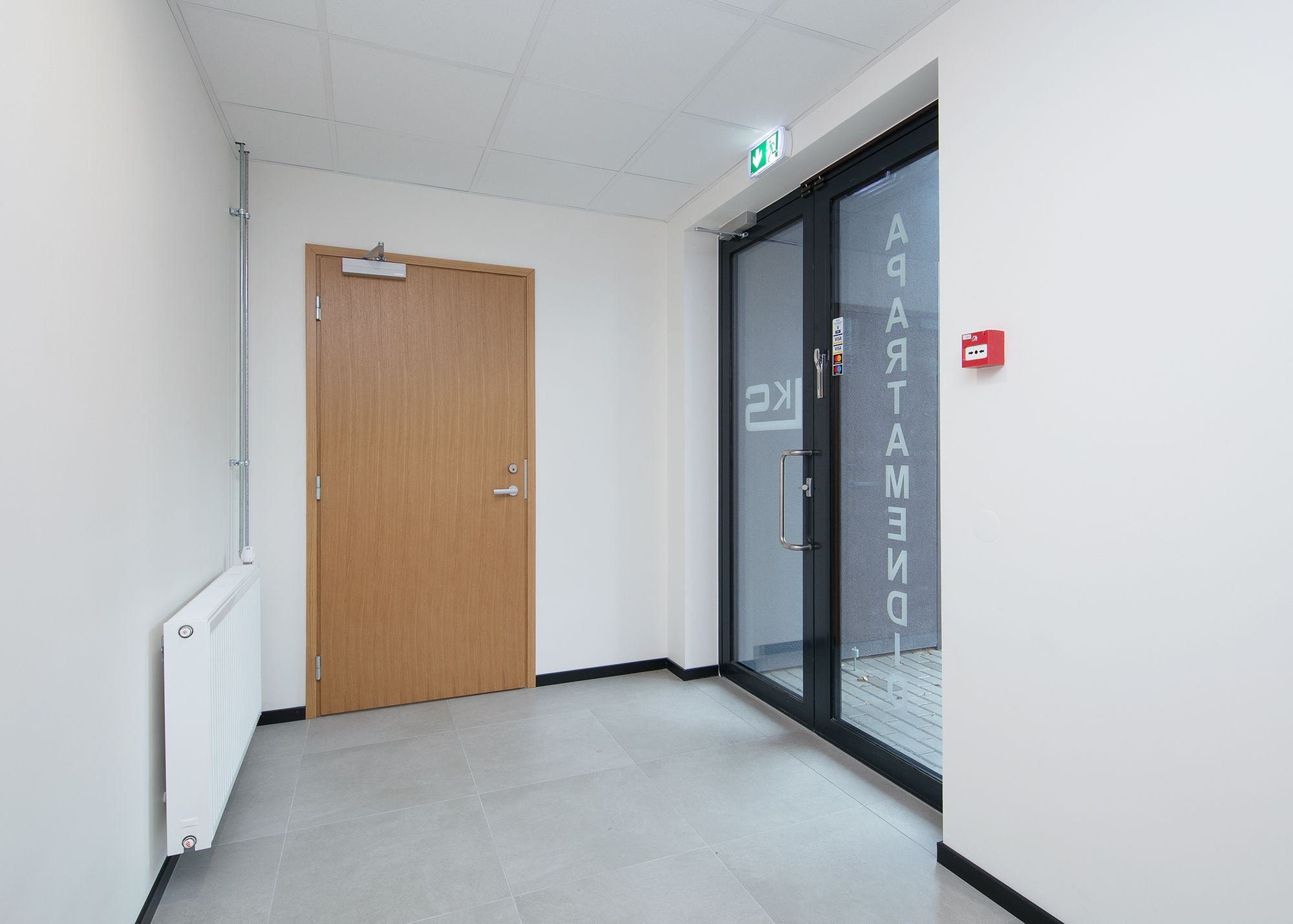A hallway with a wooden door, a glass door with a handle, and a white ceiling with a smoke detector.