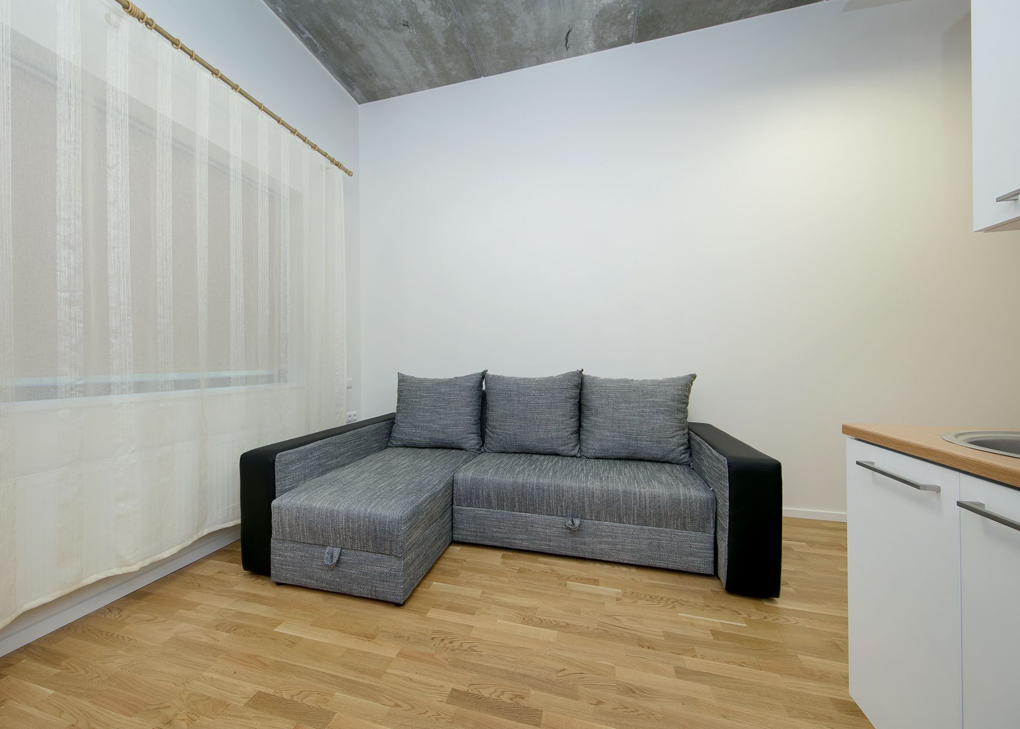 A modern room with a grey sectional sofa, a wooden floor, a white cabinet, and a window with sheer curtains.