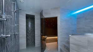 Un baño moderno con cabina de ducha de vidrio, una puerta oscura que conduce a una habitación con cama y una zona de tocador iluminada en azul.