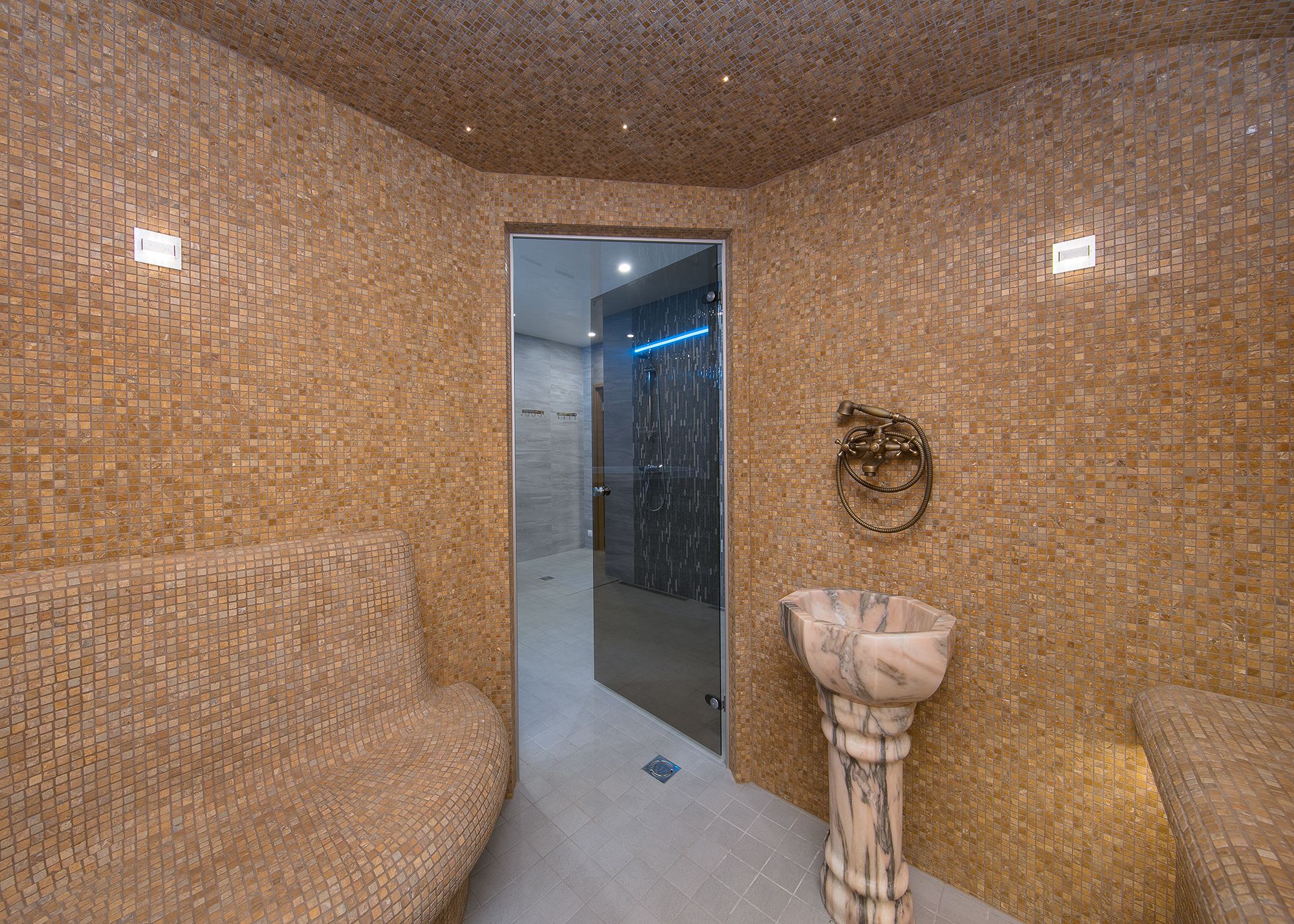 Un baño con paredes de mosaico, mampara de vidrio y lavamanos de pedestal de mármol.