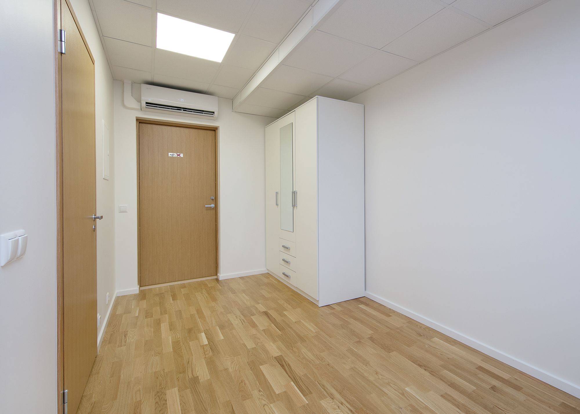 Un couloir avec un plancher en bois, une porte avec une pancarte, une armoire et un appareil de climatisation.
