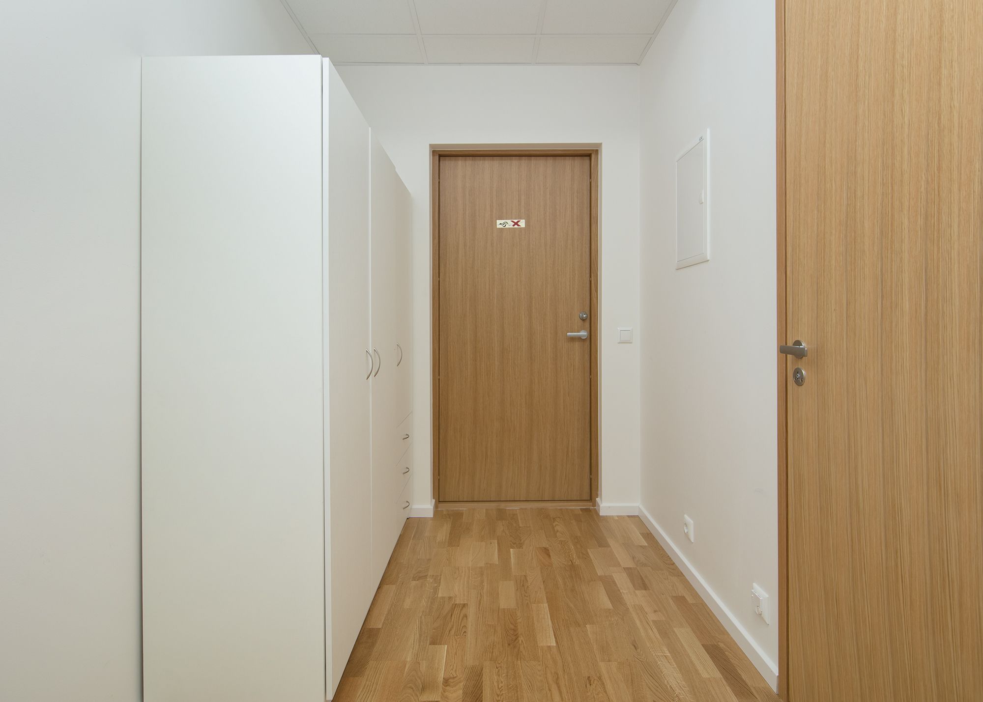 Un pasillo estrecho con suelo de madera, paredes blancas y una puerta de madera cerrada al final.