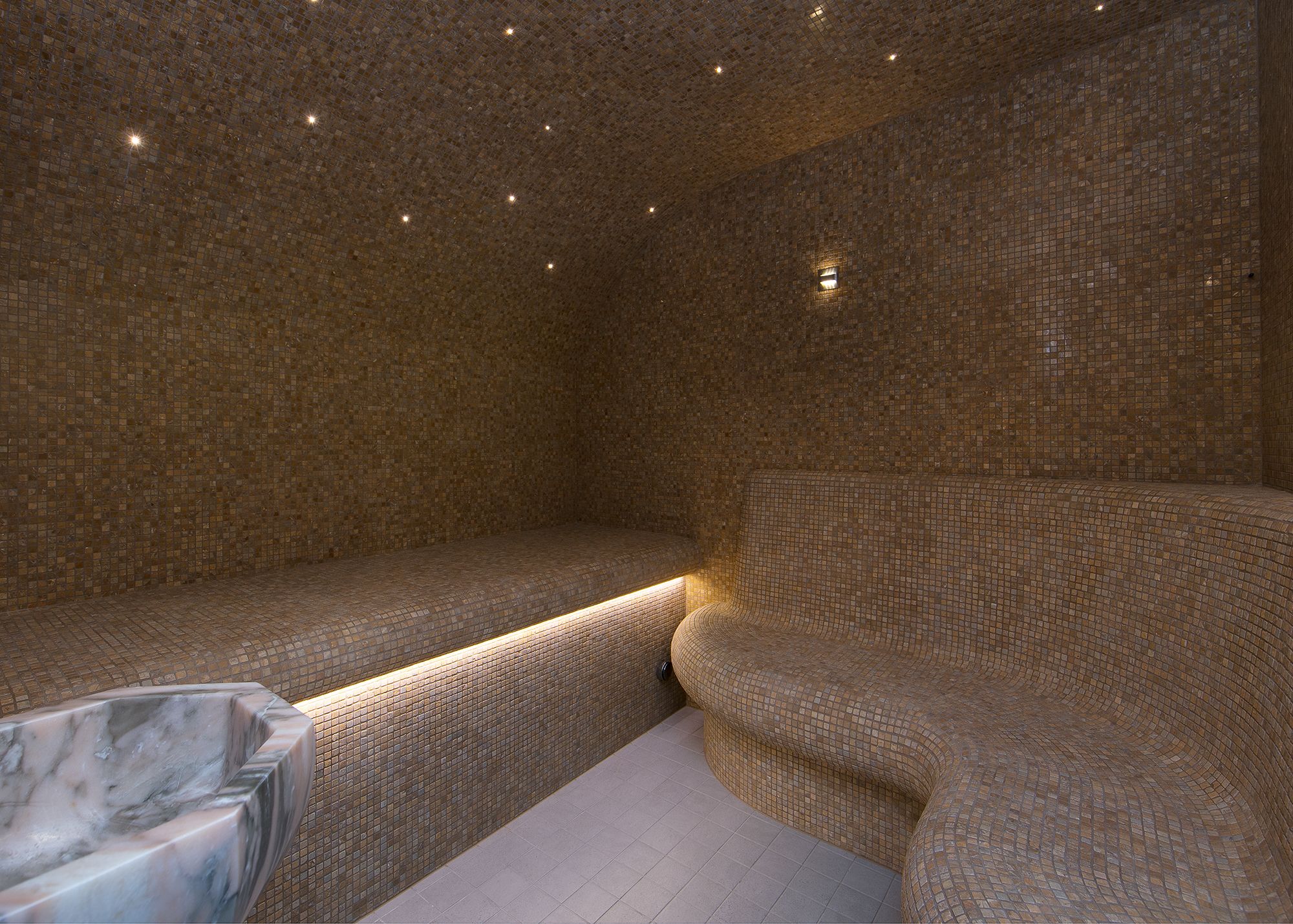A room with a mosaic-tiled wall, a marble basin, and a curved seating area.