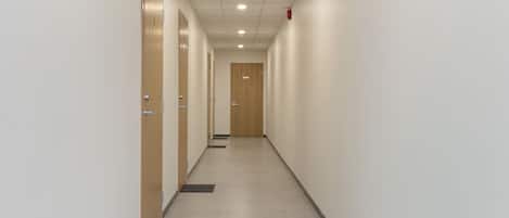A corridor with wooden doors, a tiled floor, and a fire alarm on the ceiling.