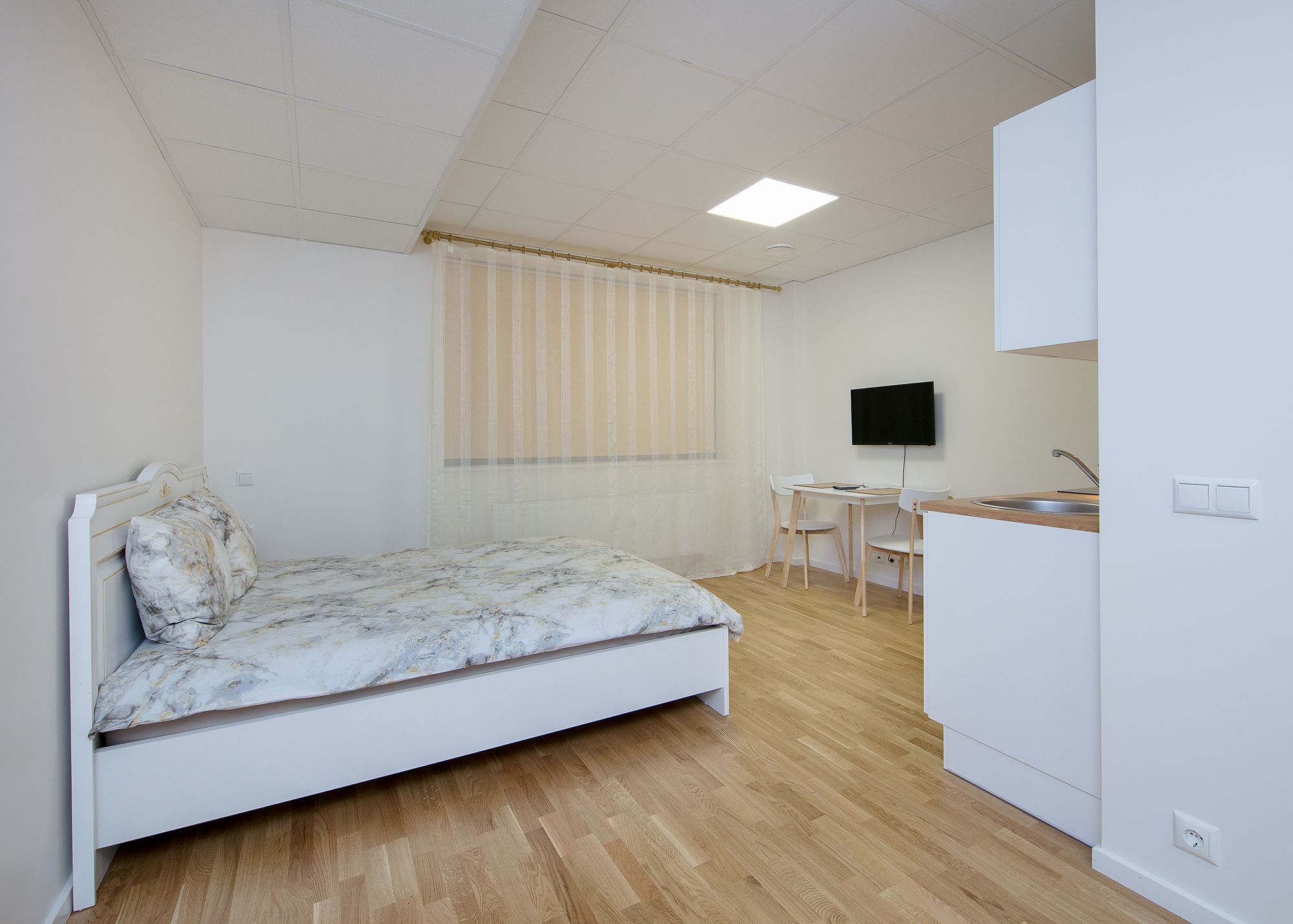 A bedroom with a bed, a desk, a chair, a TV, and a kitchenette.