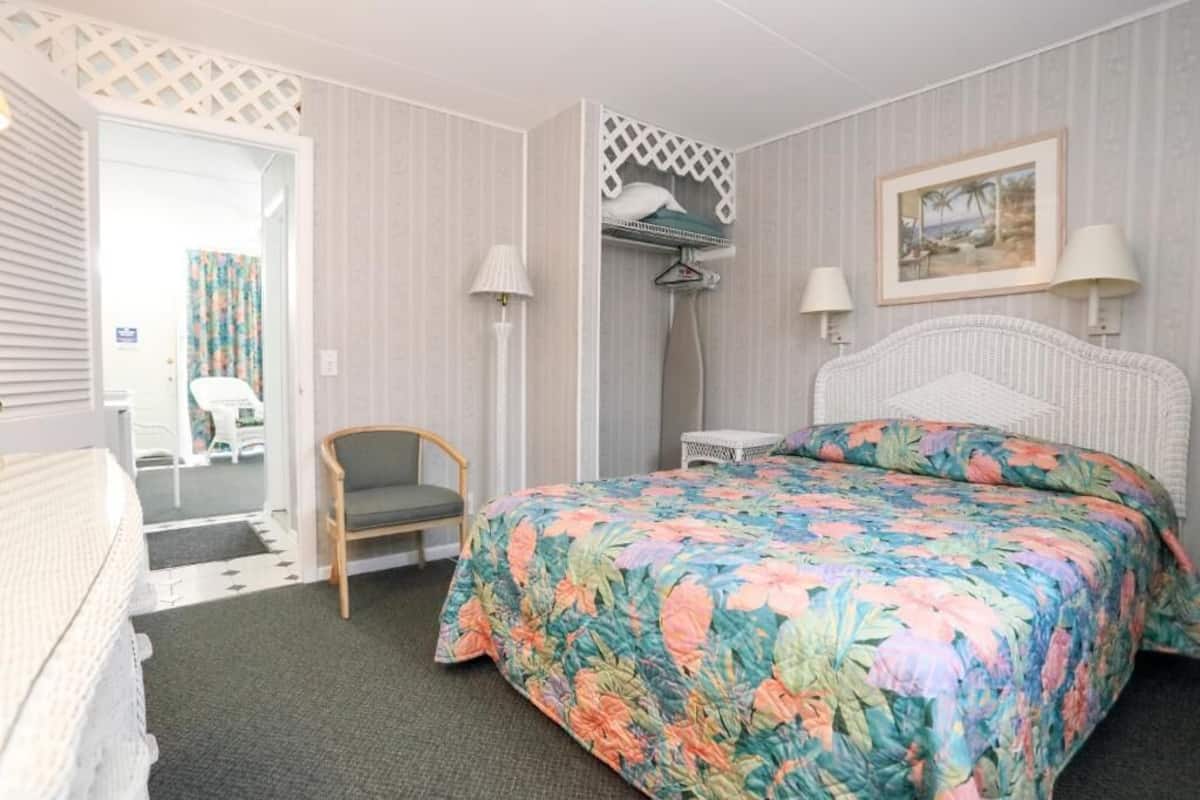 A hotel room with a bed, a chair, a small table, a hanging coat rack, and a door leading to another room.