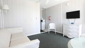 Huoneessa on valkoinen sohva, valkoinen tuoli, valkoinen lipasto, seinälle asennettu litteänäyttöinen televisio ja kattotuuletin.