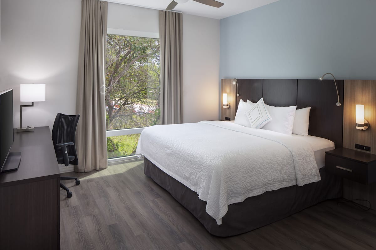 A modern hotel room with a large bed, a desk, a chair, and a window with curtains.