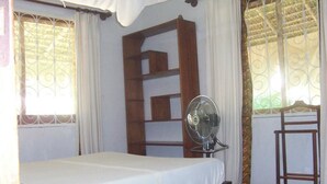 Una cama individual con colchón blanco y estructura de madera, un ventilador de pie y un pequeño estante de madera.