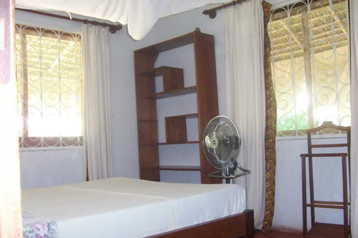 A single bed with a white mattress and a wooden frame, a standing fan, and a small wooden shelf unit.