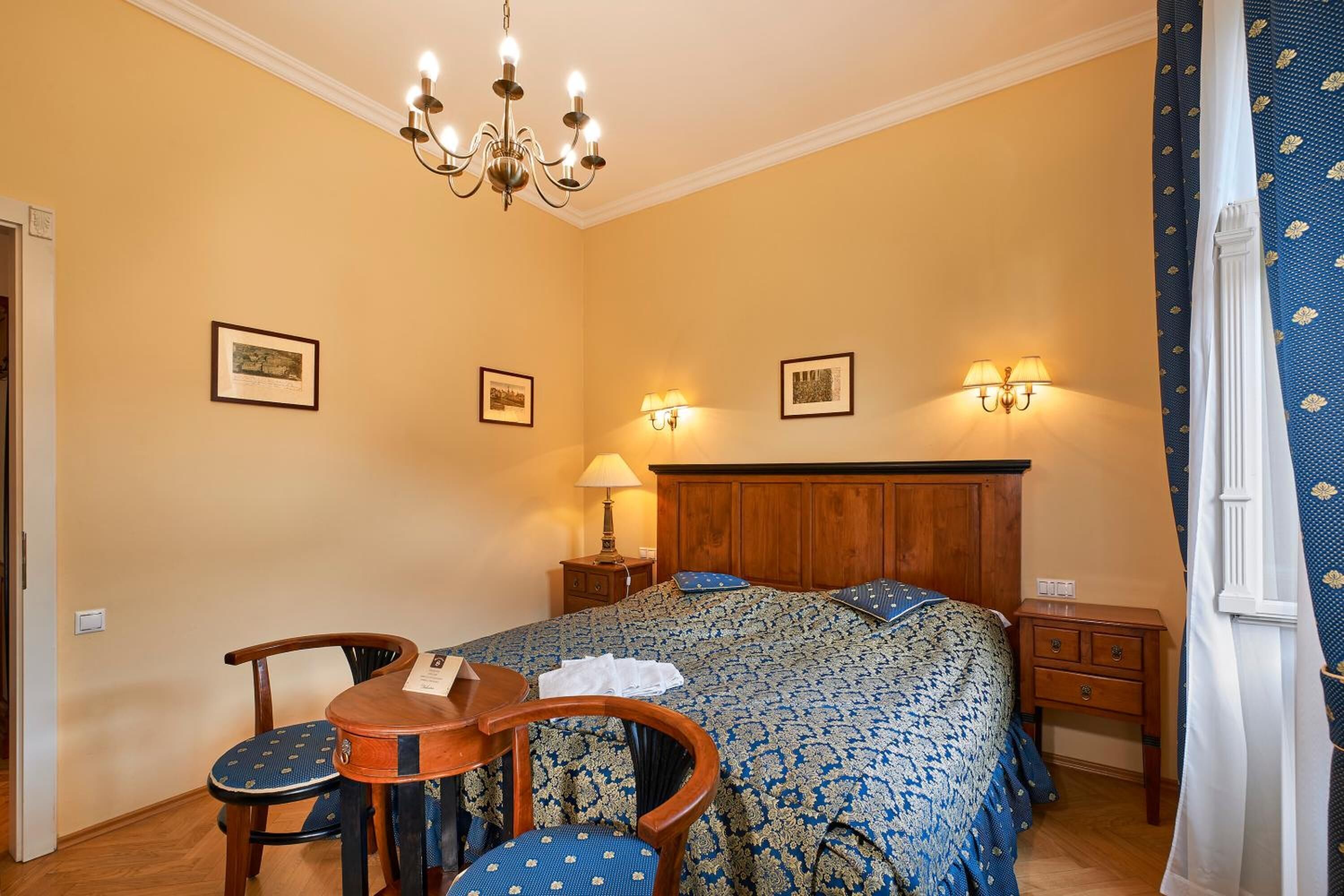 A bedroom with a wooden headboard, a bed with a patterned bedspread, a nightstand with a lamp, a wooden chair, and a small table.