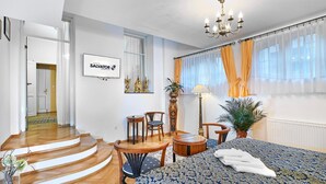 Habitación de hotel con cama, candelabro, zona de comedor con sillas y mesa, escalera y un cartel en la pared.