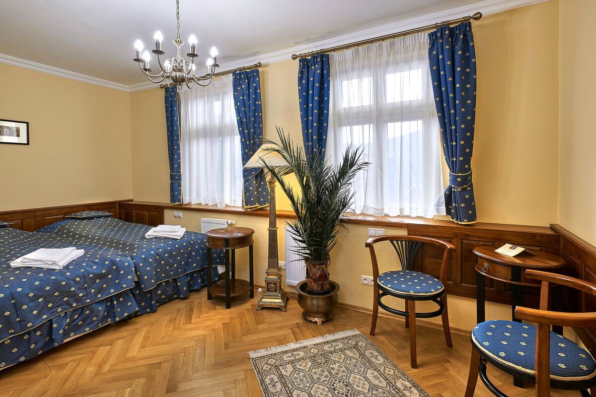 A hotel room with a large bed, two chairs, a small table, a potted plant, and a chandelier.