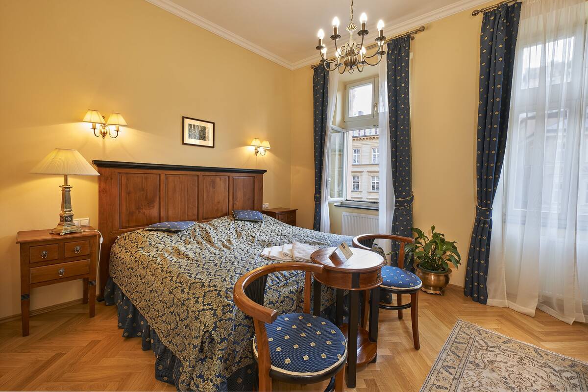 A bedroom with a bed, bedside tables, a chair, a small table, a potted plant, and a view of buildings through the window.