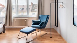Una habitación moderna con un sillón azul, un reposapiés a juego, un radiador y vistas a edificios a través de amplias ventanas.