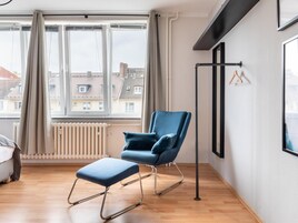 Una habitación moderna con un sillón azul, un reposapiés a juego, un radiador y vistas a edificios a través de amplias ventanas.