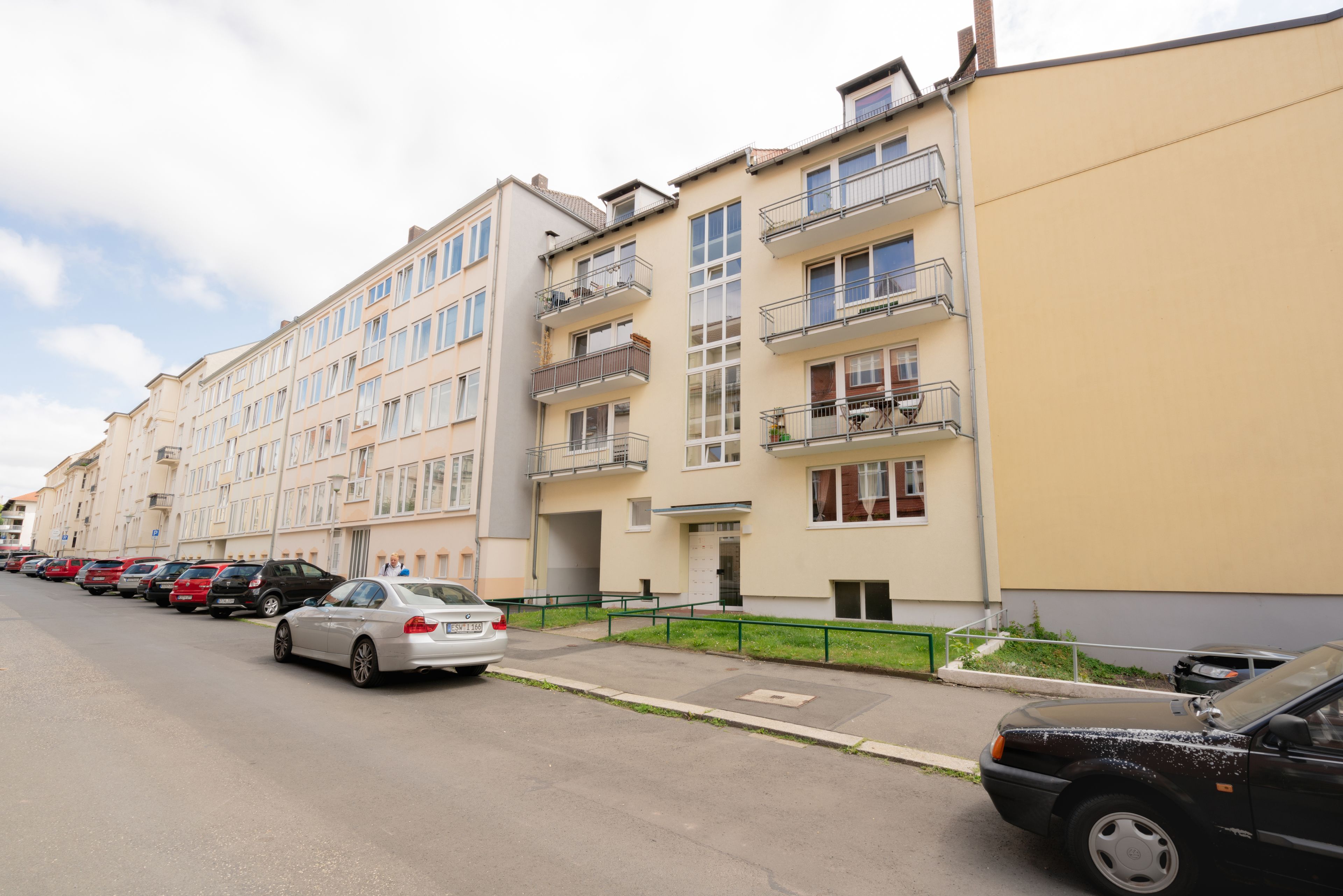 Un filare di edifici residenziali a più piani con balconi, auto parcheggiate e cielo sereno.