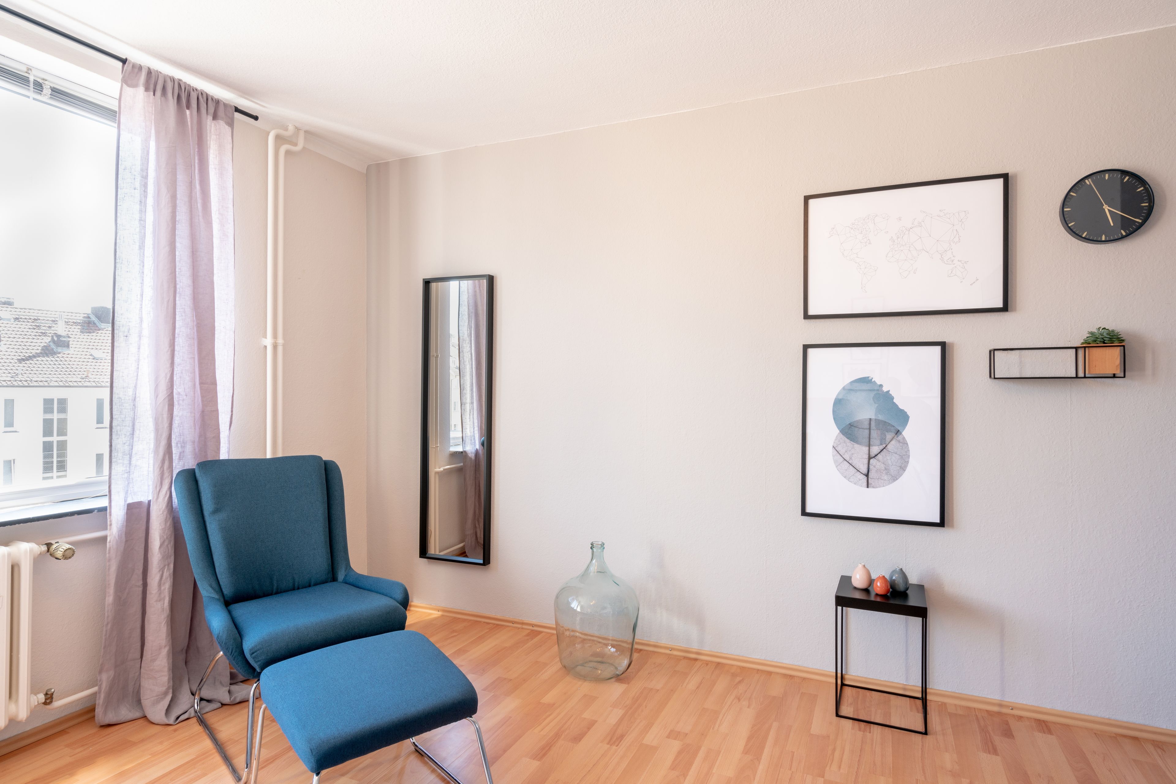 Una habitación moderna con una silla azul, un ventanal con cortinas, un mapa del mundo, un reloj y una pequeña planta en un estante.