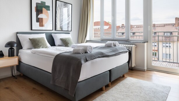 Un dormitorio moderno con un ventanal grande, una cama con ropa de cama gris y dos cuadros en la pared.