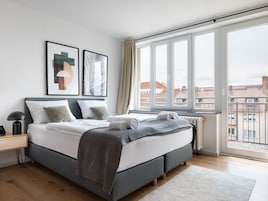 Un dormitorio moderno con un ventanal grande, una cama con ropa de cama gris y dos cuadros en la pared.