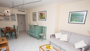 A living room with a grey sofa, a wooden coffee table with fruit, a teal cabinet, and a dining area with a table and chairs.