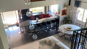 Ein geräumiger Wohnbereich mit einem weißen Sofa, einem Couchtisch und einem Esstisch mit Barhocker. Im Hintergrund ist eine Küche mit roten Schränken und einer Pendelleuchte zu sehen.