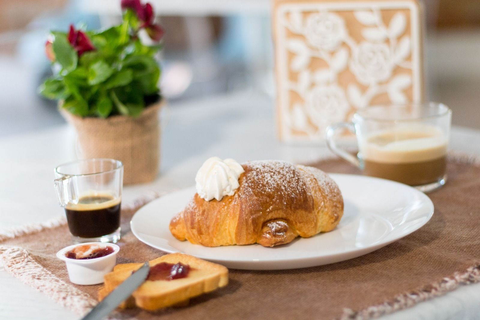 Un croissant garni de crème fouettée et saupoudré de sucre glace, un verre de café, un petit bol de confiture et une tranche de pain grillé tartinée de confiture, le tout sur une table.