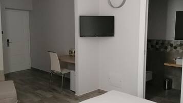 Un dormitorio con cama, escritorio, silla, televisor y baño visible a través de una puerta abierta.