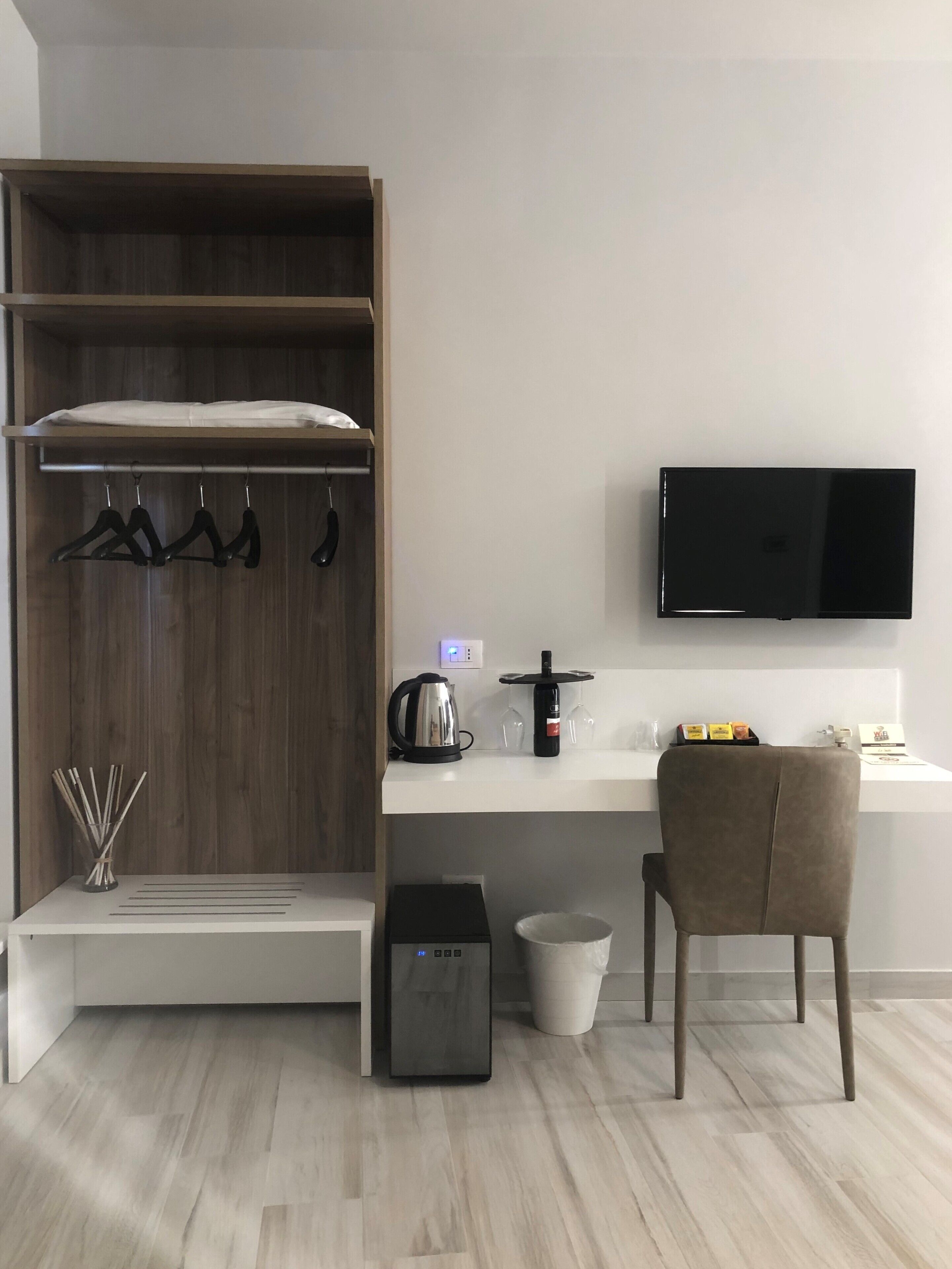 Une chambre moderne équipée d’une armoire en bois, d’un bureau blanc, d’un téléviseur à écran plat, d’une bouilloire, d’un porte-verres à vin et d’une chaise.