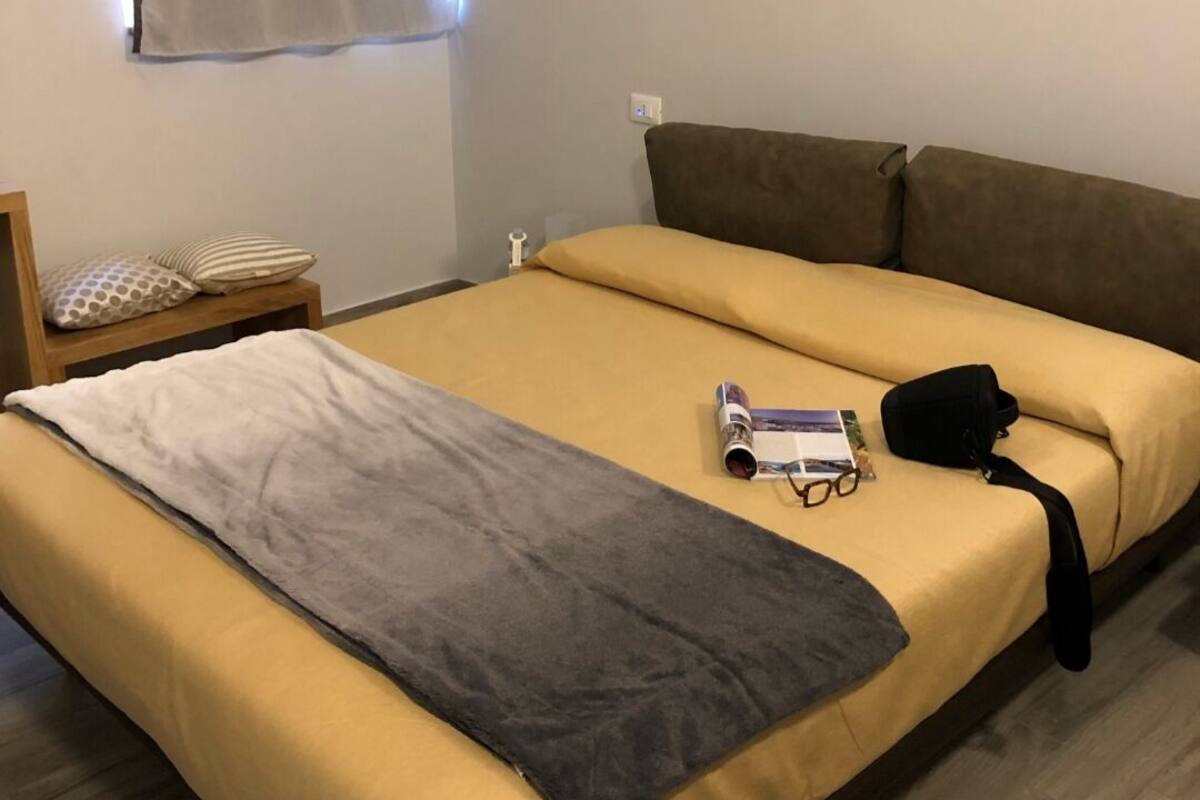 Una cama individual con colcha amarilla y manta gris, una mesita de noche de madera con teléfono, y una ventana con cortina blanca.