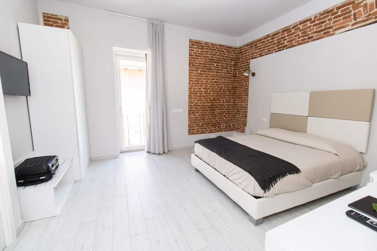 Un dormitorio con cama blanca, pared de ladrillo decorativa, armario blanco y puerta que da a un balcón.