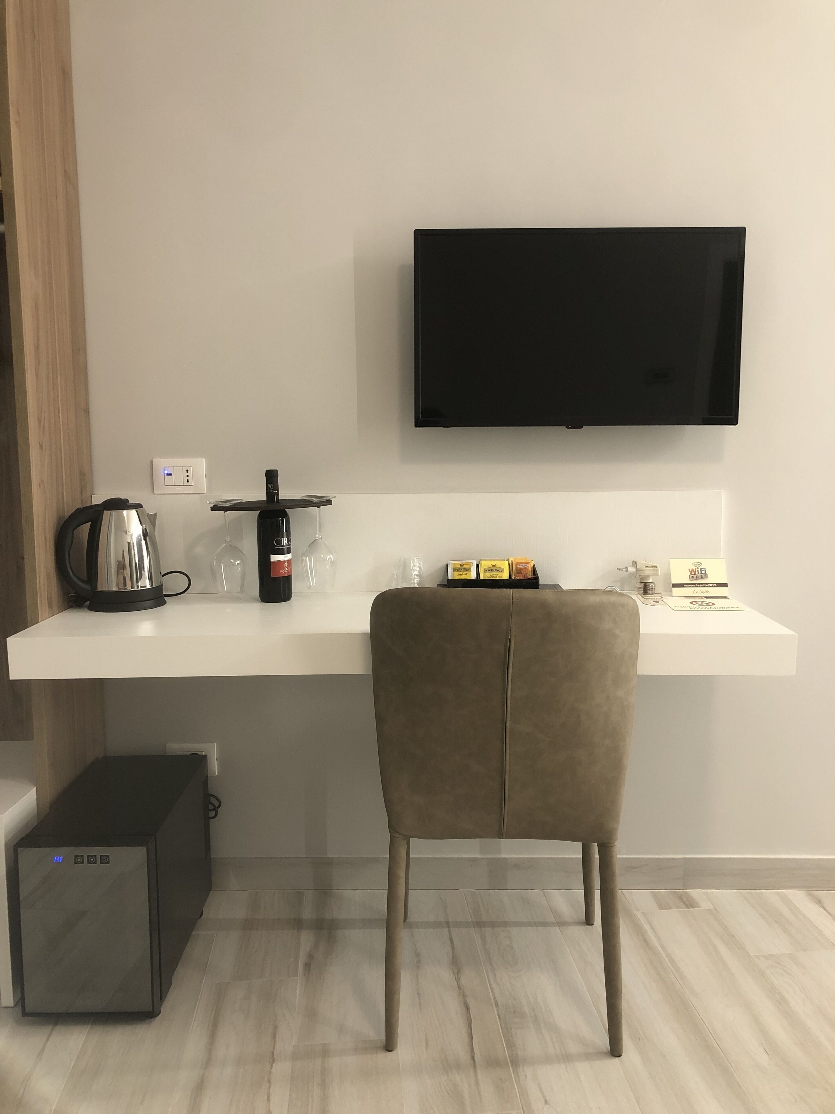 Une chambre moderne équipée d’un bureau blanc, d’un téléviseur fixé au mur, d’une bouilloire, d’un verre à vin et d’un petit réfrigérateur.