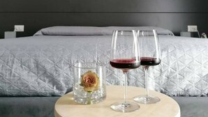Uma cama com cobertura cinza acolchoada, uma mesa de cabeceira com dois copos de vinho e um copo de água, e um vaso com uma única flor.