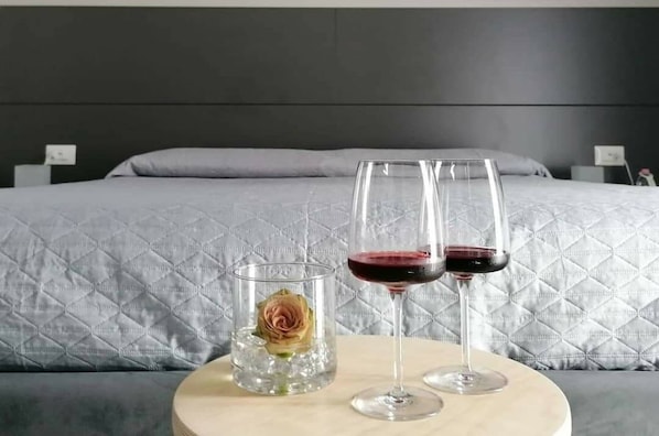 A bed with a grey quilted cover, a bedside table with two wine glasses and a glass of water, and a vase with a single flower. - Le Suite Bed & Breakfast - Holiday Home (Polistena)