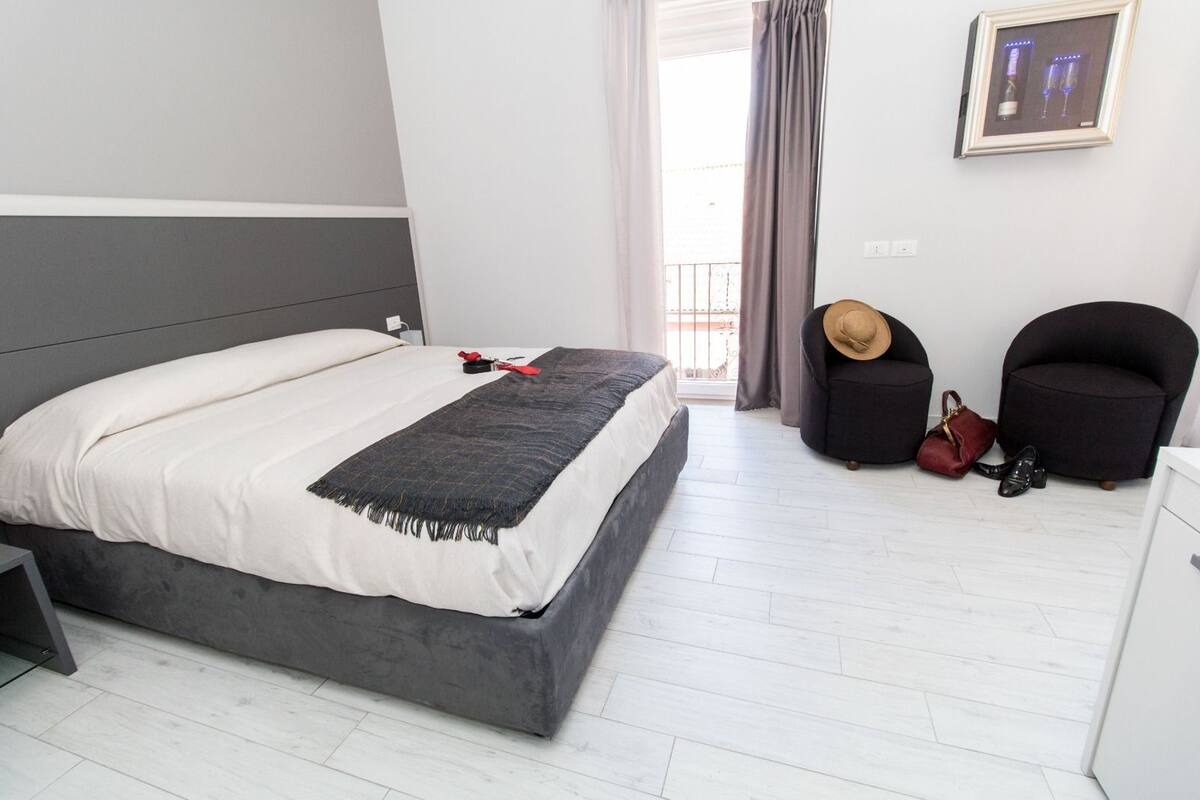 Un dormitorio moderno con una cama gris, ropa de cama blanca, un cabecero gris, una cortina gris, un sillón negro, un bolso rojo y un sombrero de madera sobre una mesita.