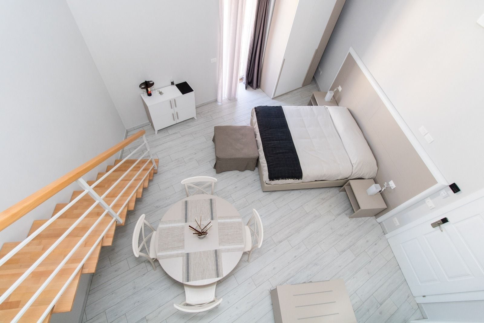 Une chambre moderne avec un lit, une table de chevet, une commode et une petite table avec des chaises. Un escalier avec des rampes en bois mène à l’étage supérieur.