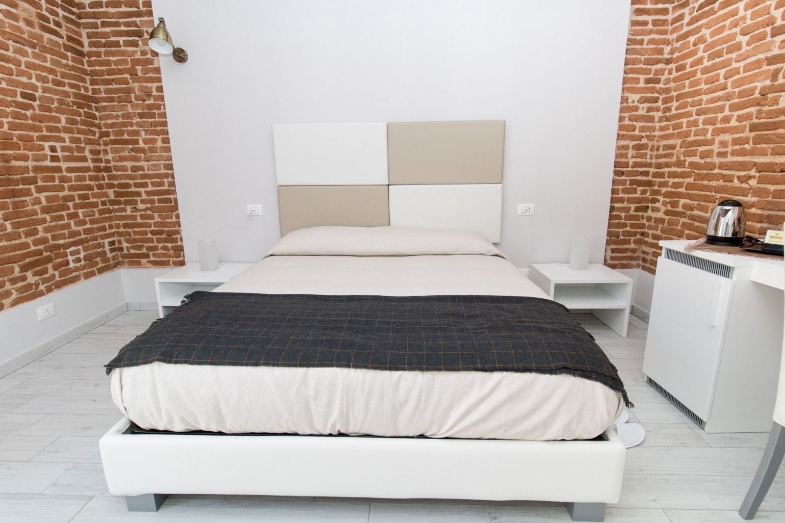 A modern bedroom with a brick wall, a white bed, a bedside table, and a small white cabinet.